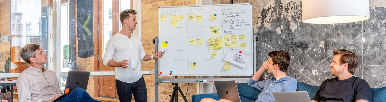 a man presenting a schedule on a white board to an audience of 3 people